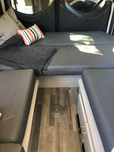 Inside a comfy **RV rental**: grey convertible seating/bed, striped pillow, blanket, and wood flooring with a table mount.
