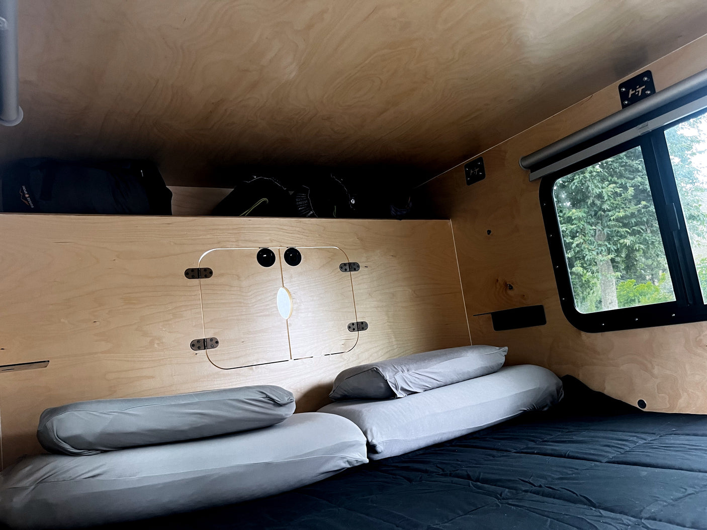 Cozy RV rental interior with wood walls, comfortable bed, overhead storage, and a window offering a lush green view outside.