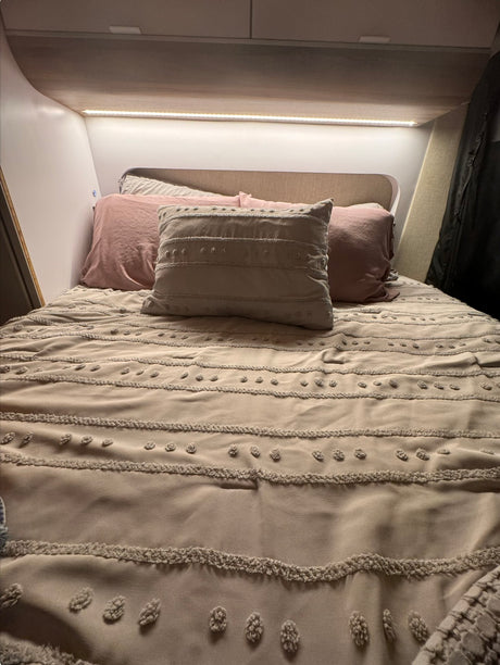 Comfortable RV rental bed features light pink and textured beige pillows on neutral bedding with strip lighting.