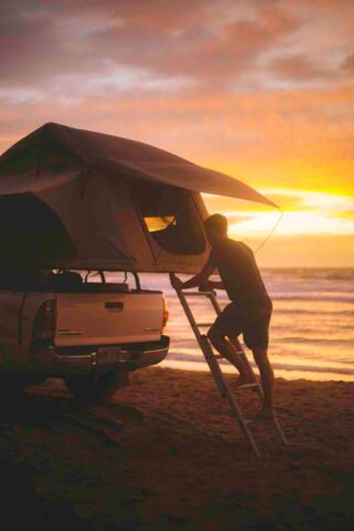 Sunset beach camping: a man climbs into a rooftop tent on a truck by the ocean. Perfect for your next RV rental adventure.