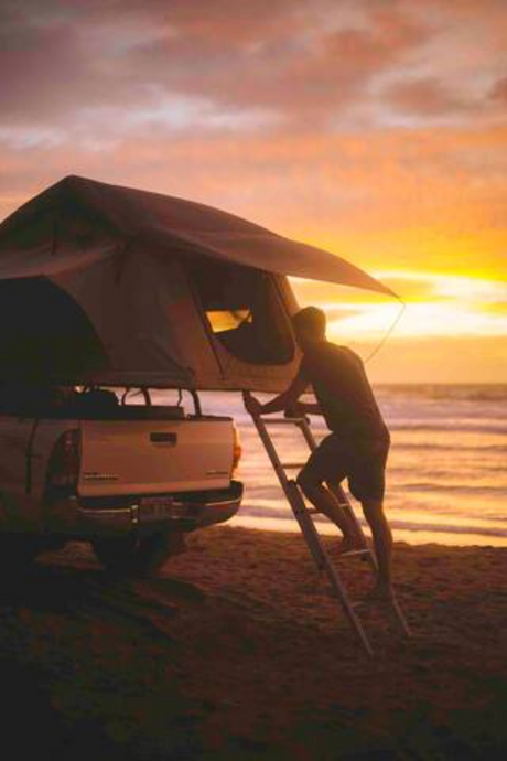 Sunset beach camping: a man climbs into a rooftop tent on a truck by the ocean. Perfect for your next RV rental adventure.