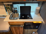 Inside an RV rental: a compact kitchen featuring a sink, two-burner stove, microwave, and refrigerator for convenient travel cooking.