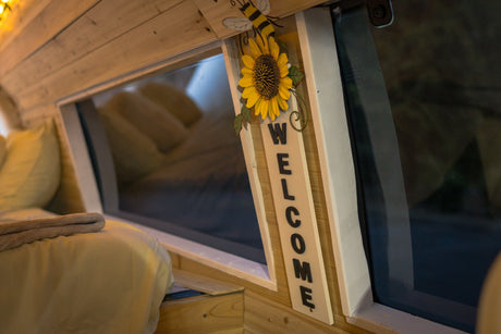 Cozy RV rental interior with wooden walls, a plush bed, and a cheerful sunflower "WELCOME" sign by the window.