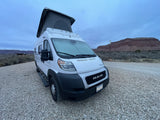 White RV rental camper van with pop-top extended, parked on gravel. Desert landscape with mesas under a cloudy sky.