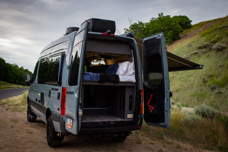Rear view of a gray camper van with open doors, revealing a cozy bed and storage. Ideal for your next RV rental adventure.