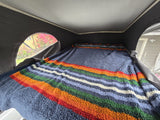 Inside a cozy RV rental pop-top, a colorful textured blanket covers the bed, with a view of blooming flowers.