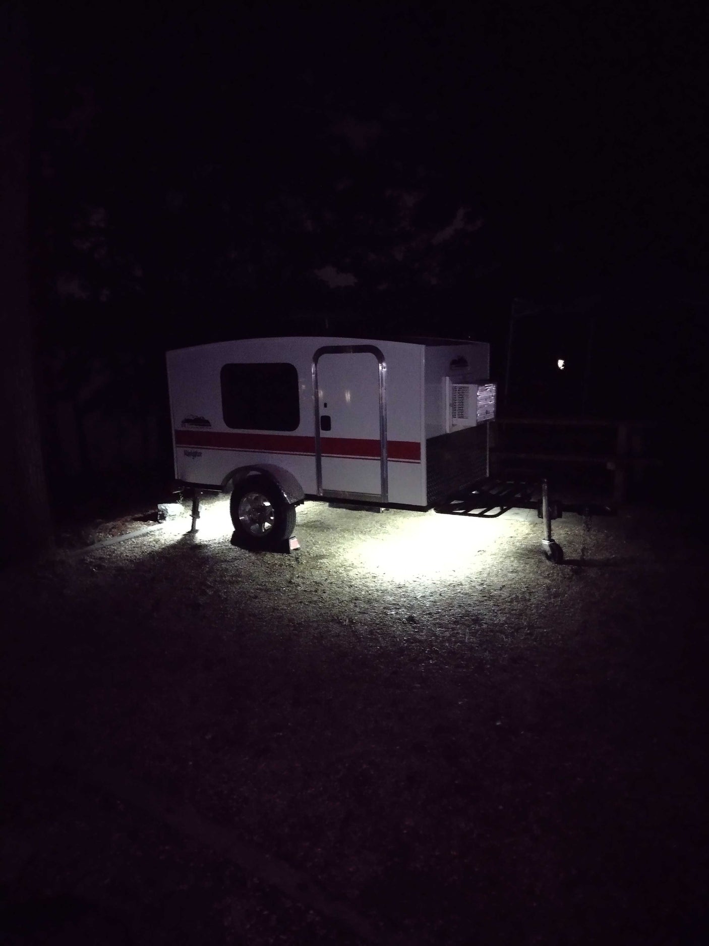 White & red compact camper RV rental, lit by underbody lights at a dark campsite. Ideal for outdoor travel.