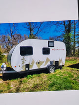 White RVE LITE teardrop RV rental featuring tree and mountain graphics, parked in a sunny outdoor setting.