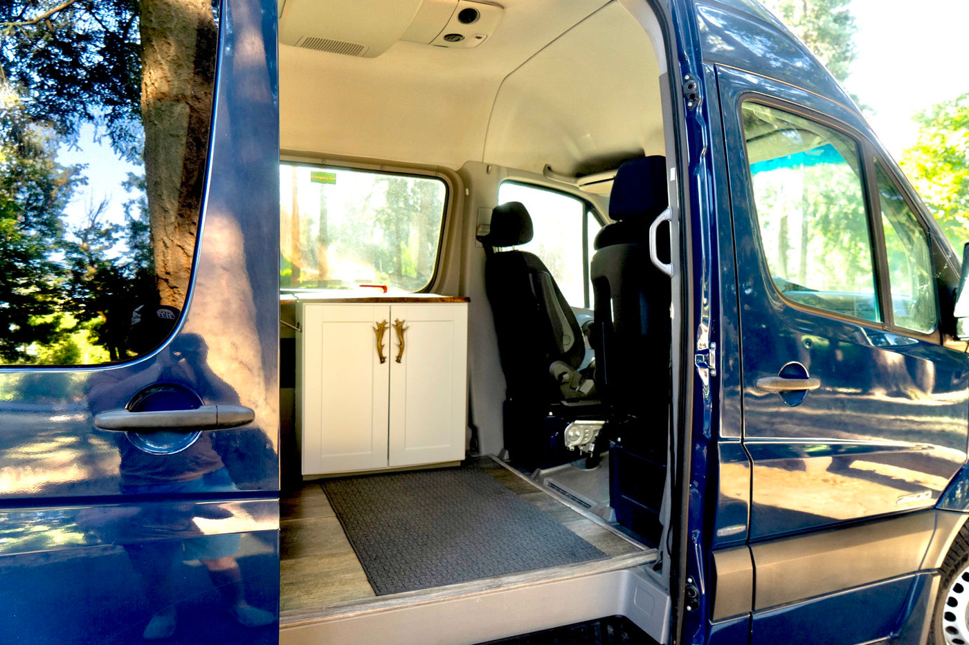  parked on a scenic dirt road with tall trees and a blue sky. Perfect for a road trip!|Blue Mercedes-Benz Sprinter RV rental parked by a river. This adventure-ready conversion van is ideal for camping & travel.|Inside view of a blue custom camper van — photo 2