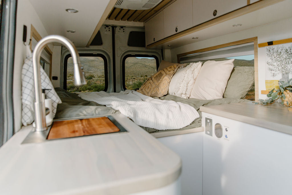 Modern RV rental interior with a cozy bed, equipped kitchen sink, & panoramic desert views through the windows.