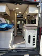 Step inside this modern RV rental, showcasing a functional and comfortable interior with a bed, kitchenette, and aisle space.