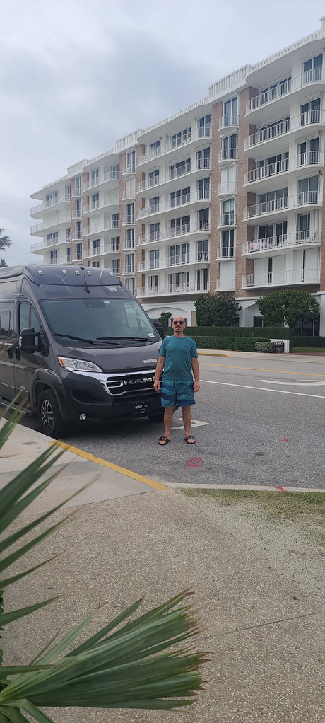 Man stands next to his gray RV rental van, a Winnebago, parked on a street in front of a grand building.