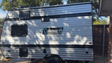 A clean silver Coleman travel trailer, a great rv rental option, with black trim, windows, and vents, parked outdoors.
