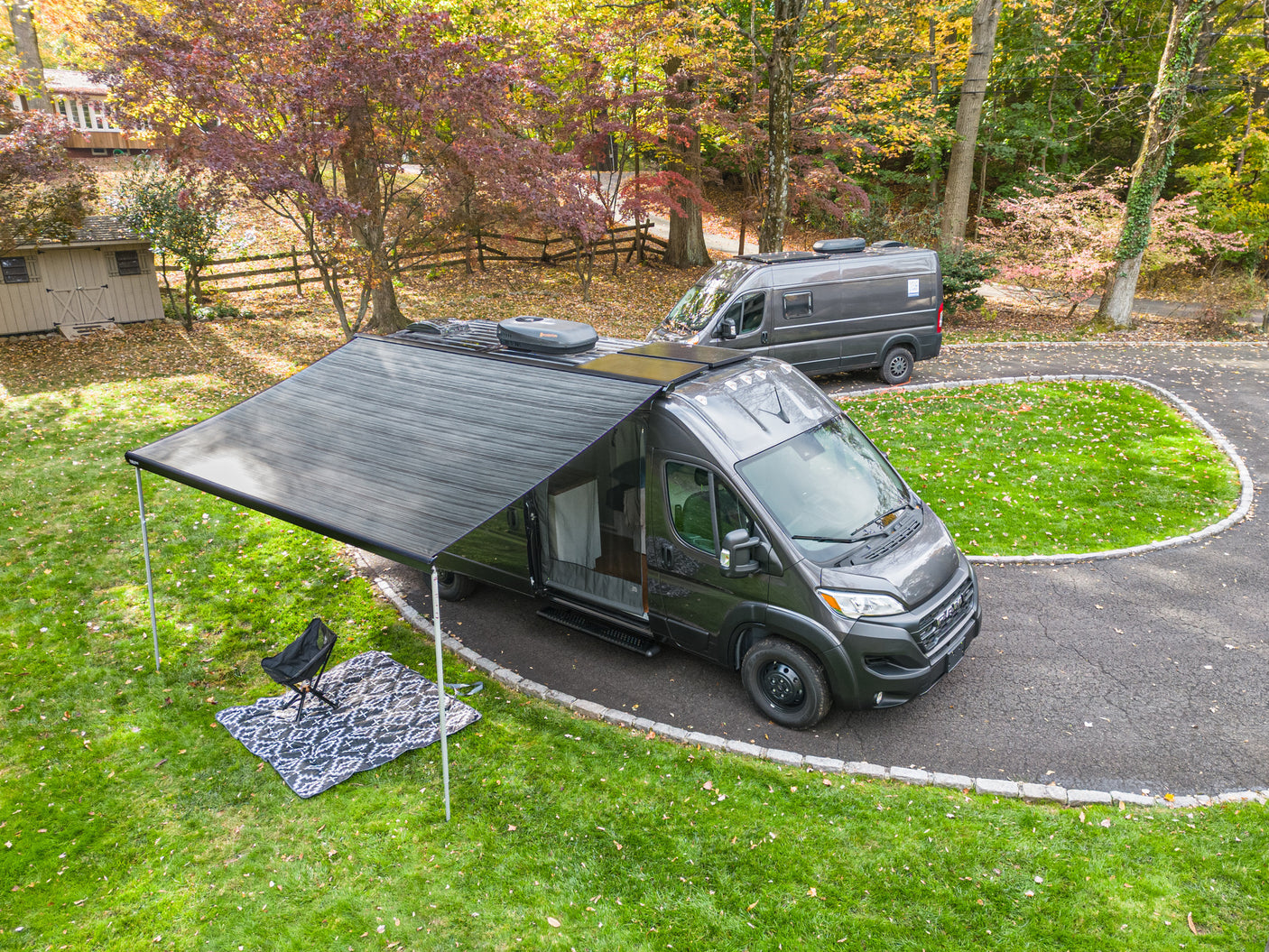 Experience adventure with an RV rental! Two dark gray campervans on a driveway, one with an extended awning, chair & blanket.