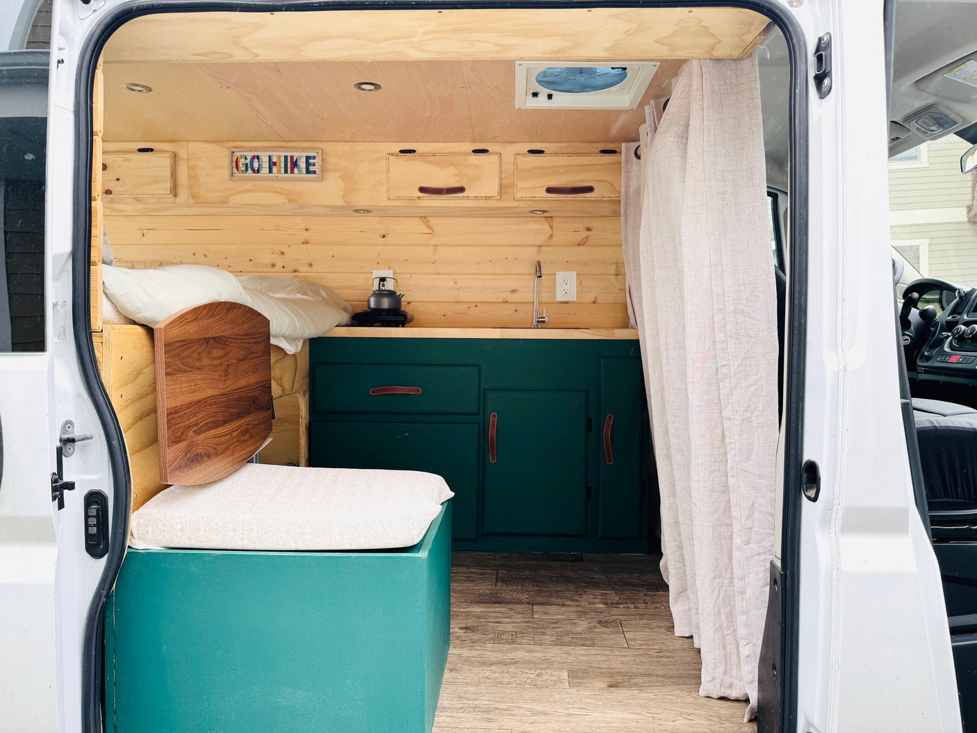 Explore this charming RV rental! A custom camper van interior features a comfy bed, dark green kitchen, and wood accents.