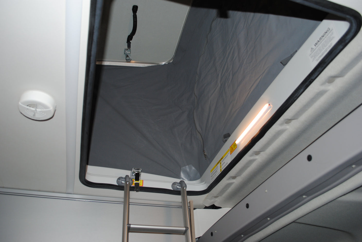 An RV rental's pop-top sleeping loft, accessed by a ladder, features a warning label & bright light.