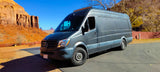 Dark grey Mercedes Sprinter RV rental parked at Monument Valley with stunning red rock mesas under a clear blue sky. Travel adventure.