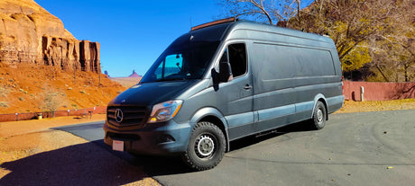 Dark grey Mercedes Sprinter RV rental parked at Monument Valley with stunning red rock mesas under a clear blue sky. Travel adventure.