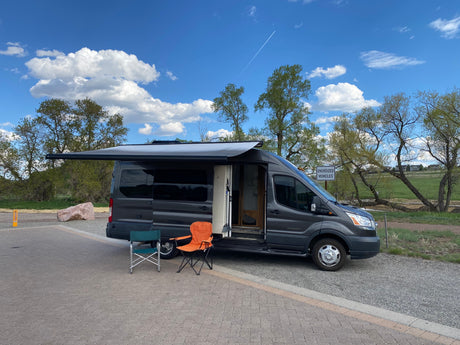 A dark grey RV rental van, with its side door open and awning extended, parked on pavers with two chairs nearby. Trees and sky in background.