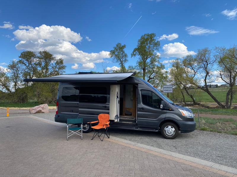 A dark grey RV rental van, with its side door open and awning extended, parked on pavers with two chairs nearby. Trees and sky in background. — photo 3