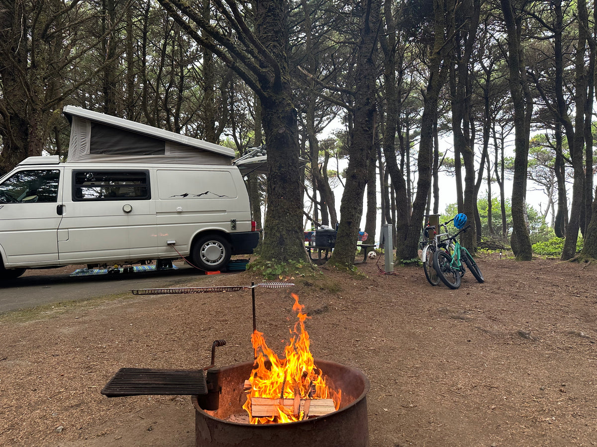 Cozy RV rental campsite in a dense forest with a bright campfire, van, and bicycles for outdoor adventure. — photo 9