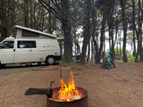 Cozy RV rental campsite in a dense forest with a bright campfire, van, and bicycles for outdoor adventure.