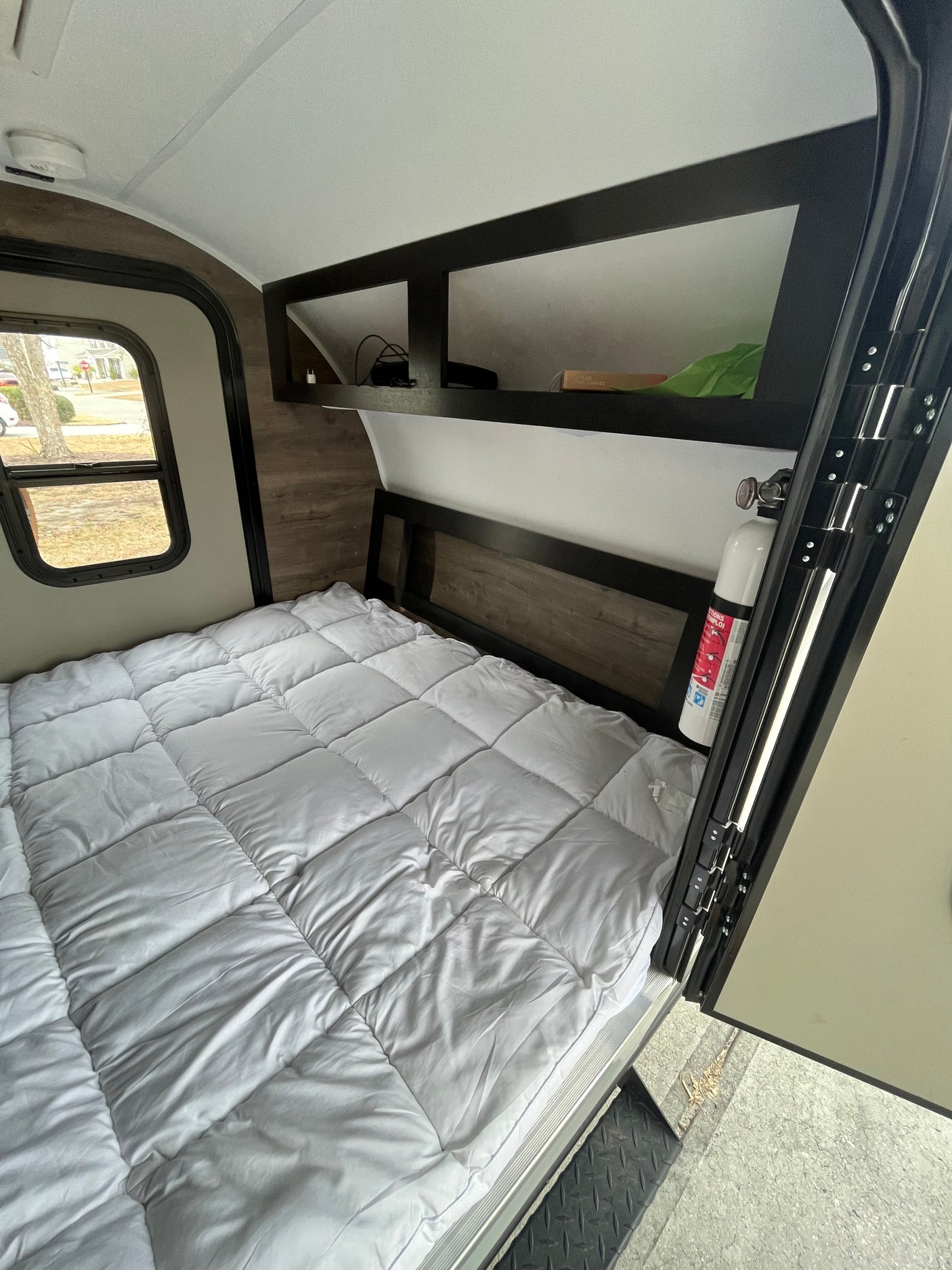 Inside an rv rental: a compact bedroom with a white quilted bed, overhead shelves, window, and fire extinguisher.