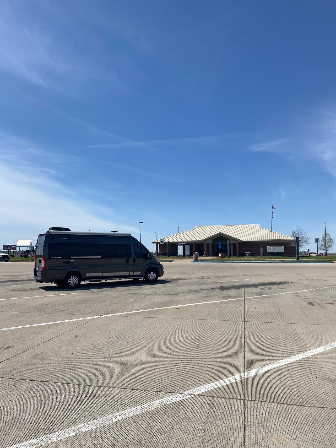 A dark grey RV rental, a Travato camper van, is parked in a vast concrete lot at a sunny rest stop under a blue sky.