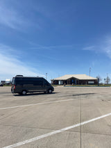 A dark grey RV rental, a Travato camper van, is parked in a vast concrete lot at a sunny rest stop under a blue sky.