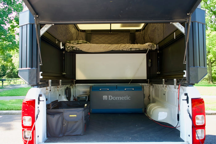 Inside a white pickup truck's open camper, showing a bed, Dometic fridge, & storage. Ideal for an RV rental camping adventure.
