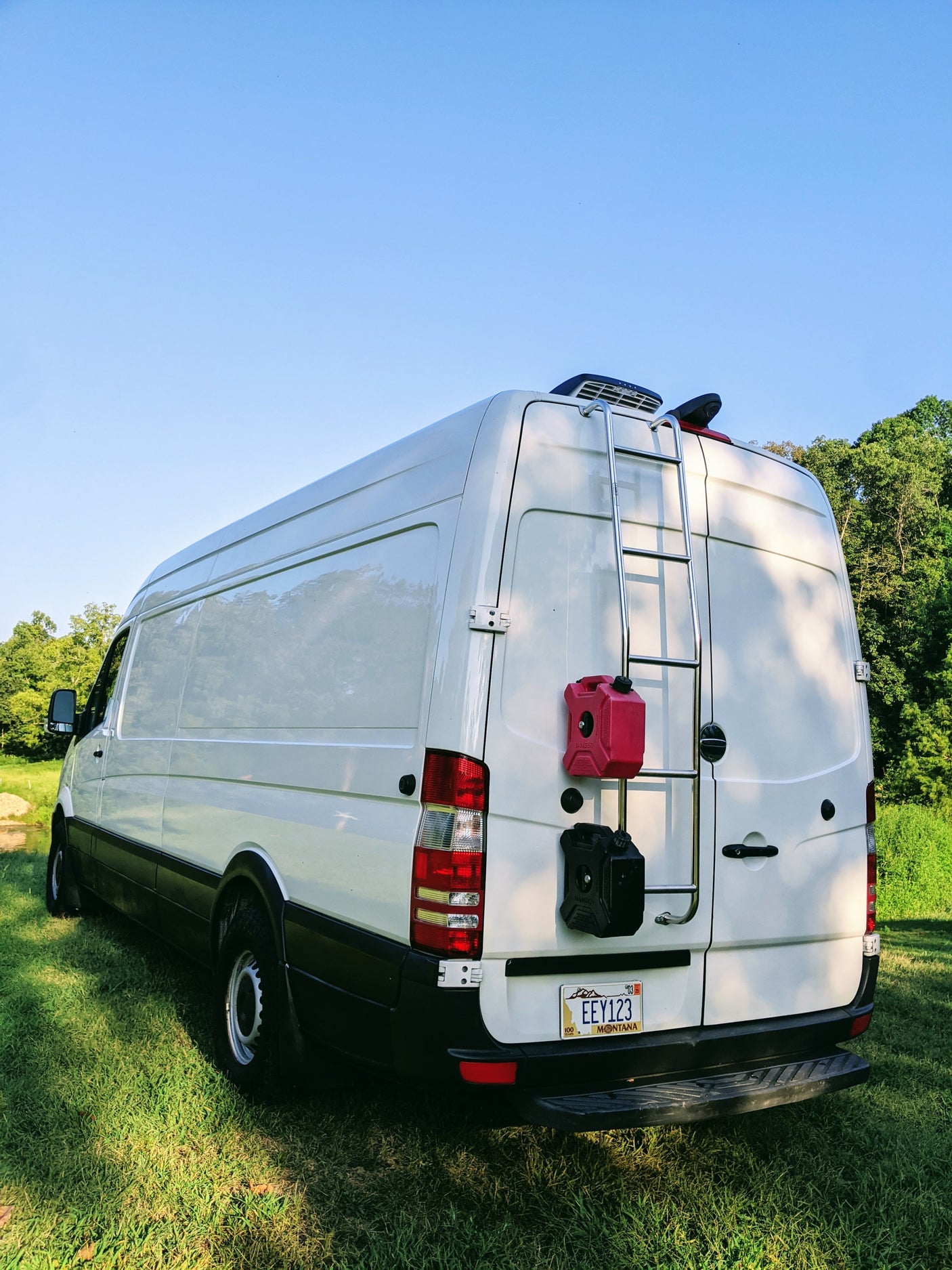 White camper van with a rear ladder, fuel containers, and Montana license plate on grass. Ideal for rv rental.
