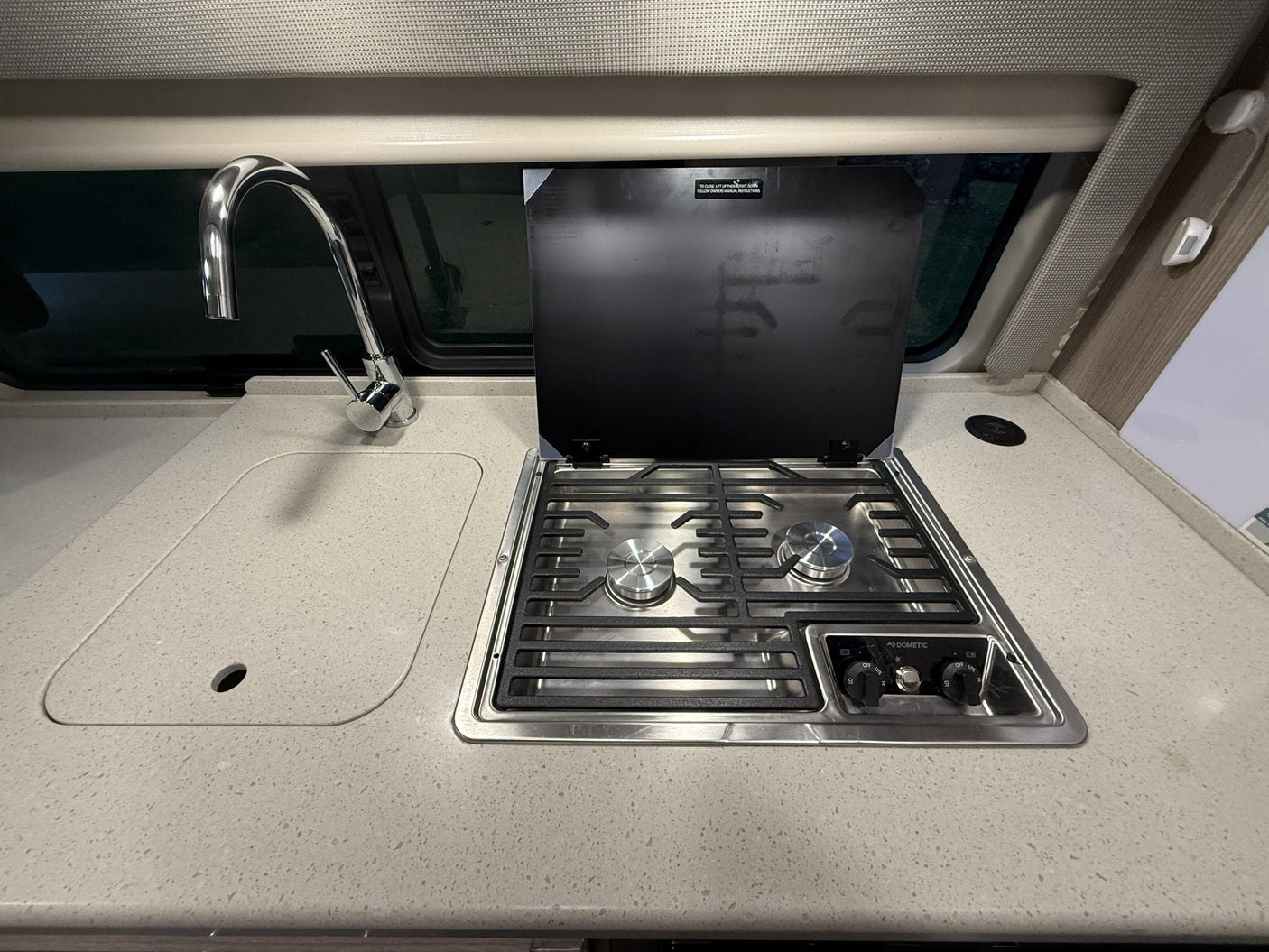 Compact RV rental kitchen featuring a shiny faucet over a sink and a Dometic two-burner stovetop with its protective lid open.