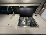 Compact RV rental kitchen featuring a shiny faucet over a sink and a Dometic two-burner stovetop with its protective lid open.