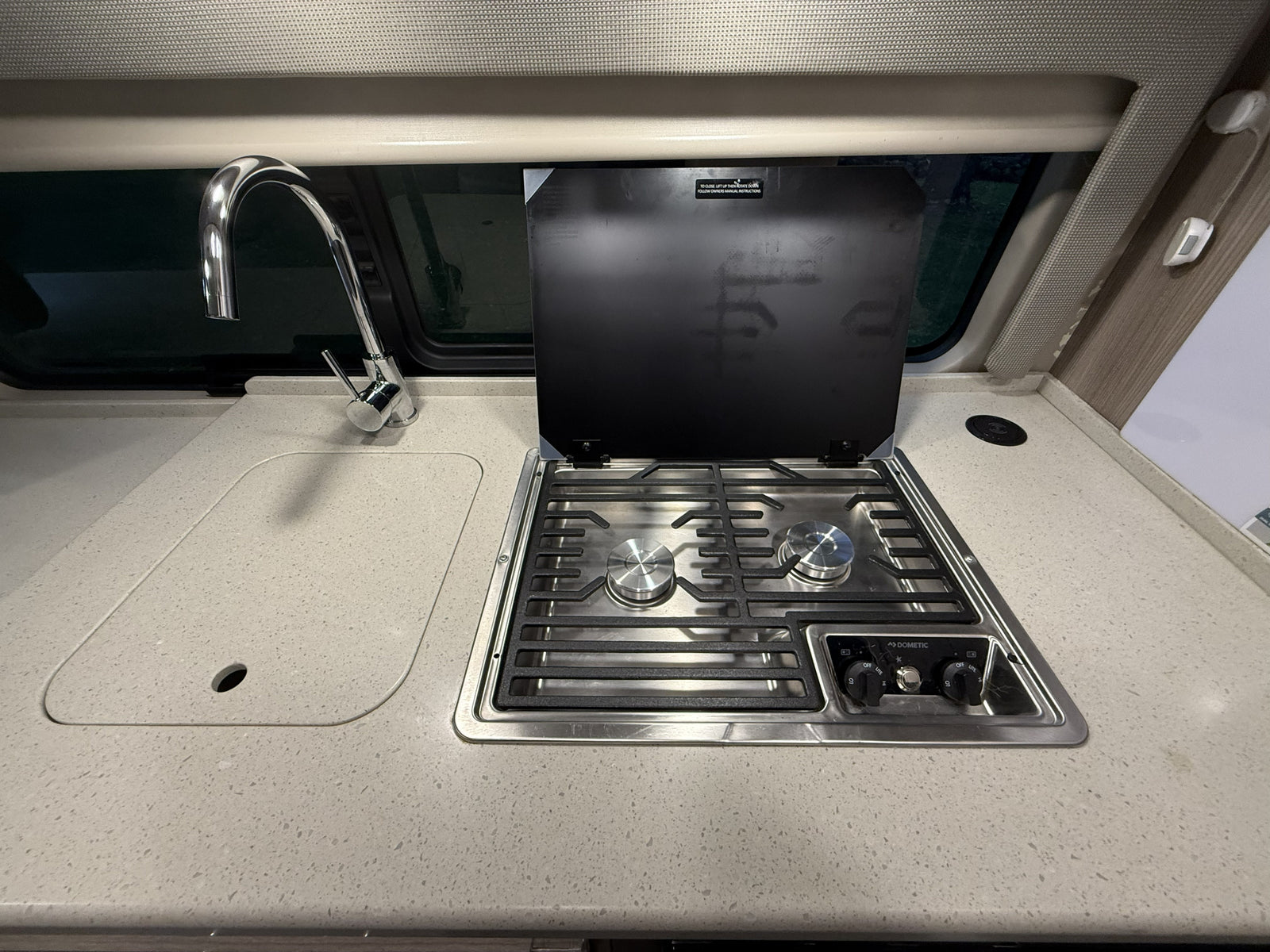 Compact RV rental kitchen featuring a shiny faucet over a sink and a Dometic two-burner stovetop with its protective lid open.