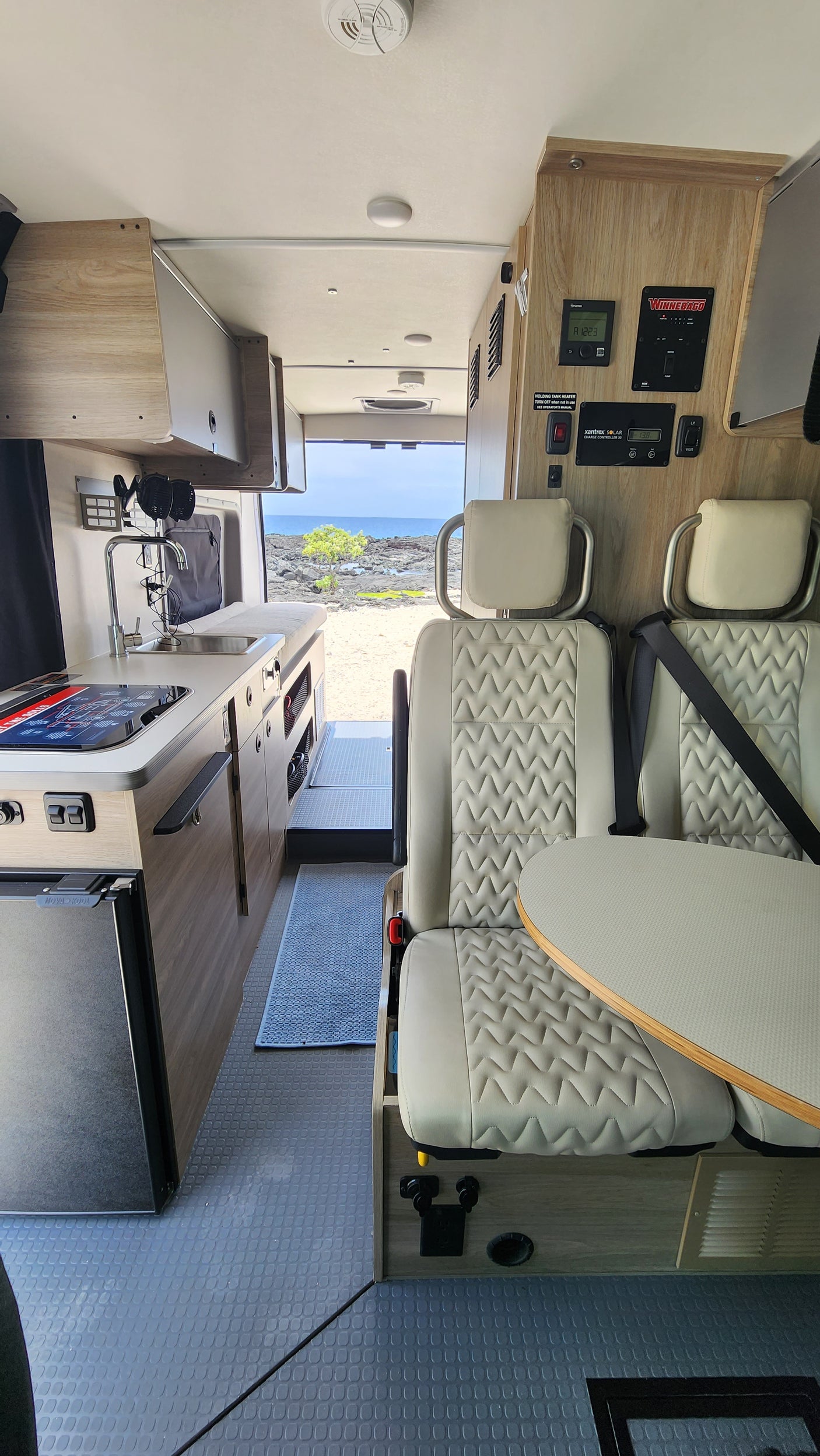 Modern rv rental interior: compact kitchen, seating with table, and control panels. A scenic ocean view is visible out the back.