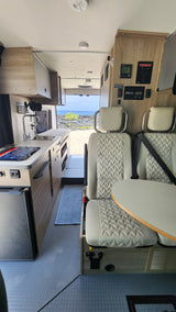 Modern rv rental interior: compact kitchen, seating with table, and control panels. A scenic ocean view is visible out the back.