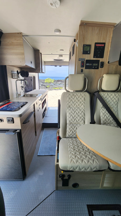 Modern rv rental interior: compact kitchen, seating with table, and control panels. A scenic ocean view is visible out the back.