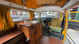 Interior of a vintage RV rental camper van with kitchenette, plaid passenger seat, and wood flooring.