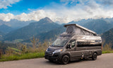 Modern grey camper van (RV rental) with pop-top tent on a scenic mountain road, perfect for nature adventures.