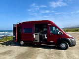 Red RV rental camper van, doors open, revealing its interior & ocean view, parked on a dirt path for travel adventure.