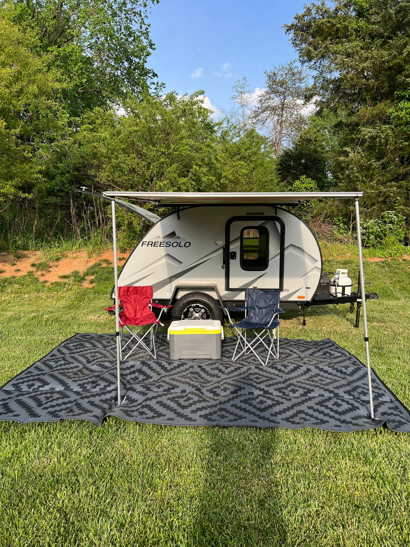Relaxing outdoor RV rental setup: a light grey teardrop camper with an extended awning, chairs, and cooler on a patterned mat.