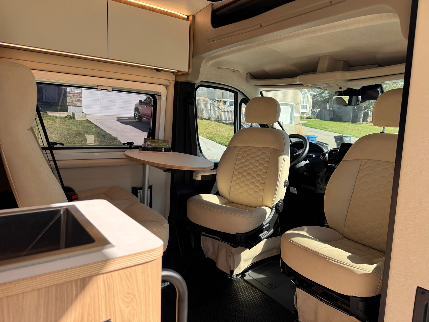 Bright RV rental interior features beige swivel front seats, a small dining table with books, and sleek overhead cabinets. A sink is on the left. — photo 8