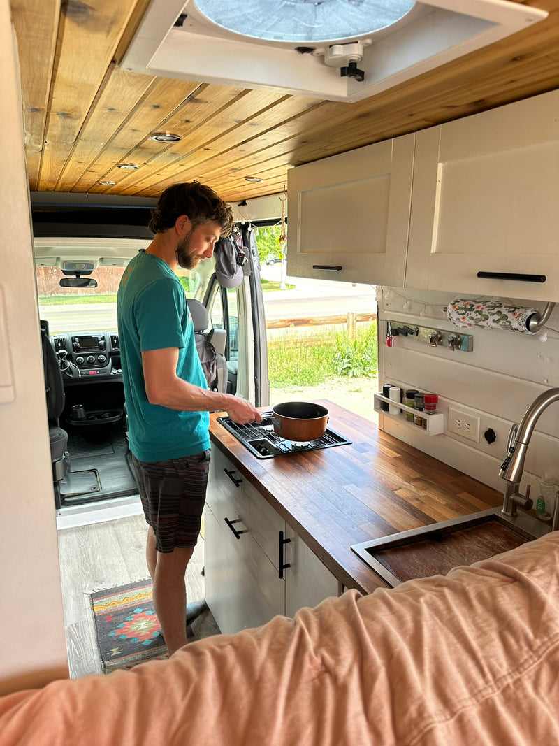 Man cooks in a modern campervan kitchen, great for an rv rental. Features wood ceiling, white cabinets, & stove. — photo 10