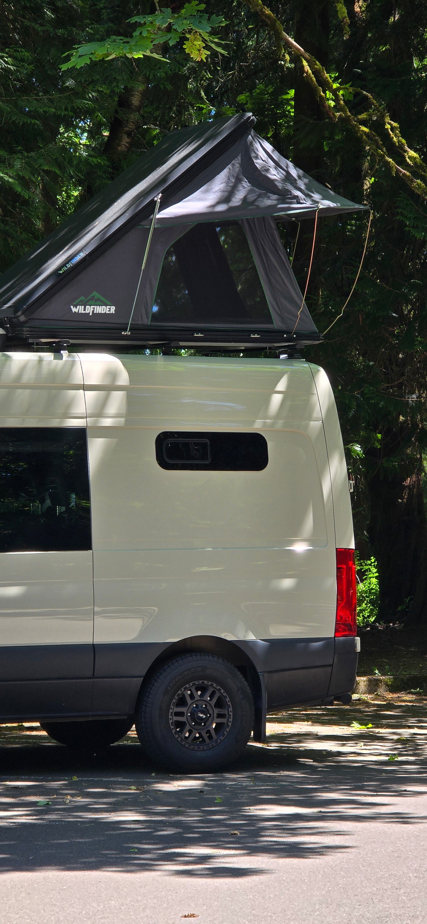 Light-colored RV rental van featuring a Wildfinder rooftop tent, parked in a lush, tree-filled camping spot.