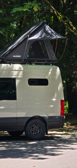 Light-colored RV rental van featuring a Wildfinder rooftop tent, parked in a lush, tree-filled camping spot.