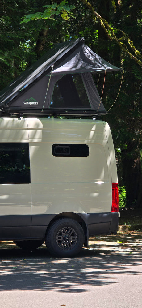 Light-colored RV rental van featuring a Wildfinder rooftop tent, parked in a lush, tree-filled camping spot.