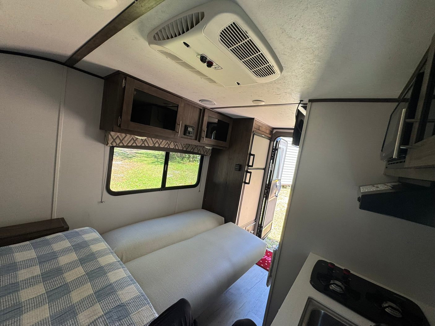 RV rental interior: bright living area with cushioned seating, a checkered blanket, compact kitchen, and overhead AC unit.