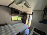 RV rental interior: bright living area with cushioned seating, a checkered blanket, compact kitchen, and overhead AC unit.