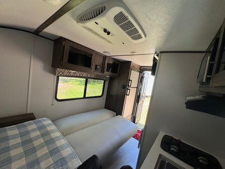 RV rental interior: bright living area with cushioned seating, a checkered blanket, compact kitchen, and overhead AC unit.