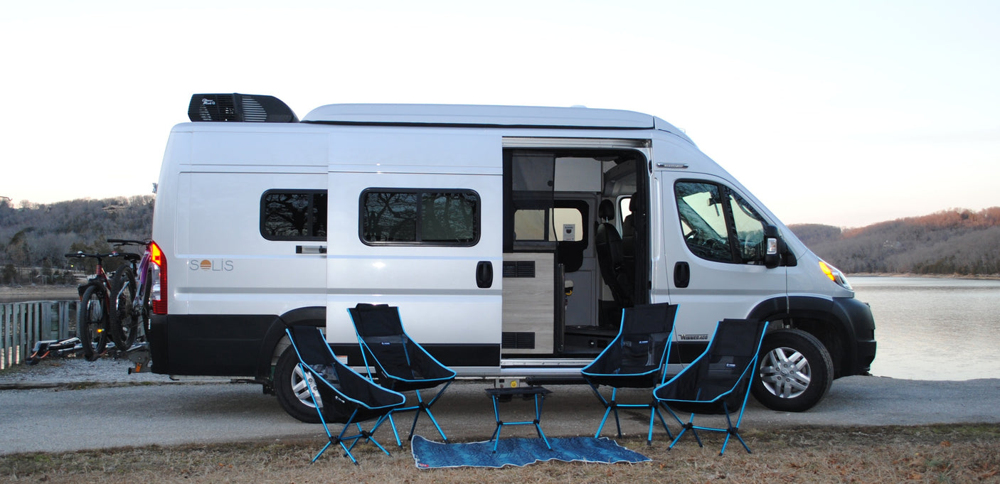 A bright Winnebago Solis RV rental parked by a lake with bikes on the rear. Camping chairs & a mat are set out.